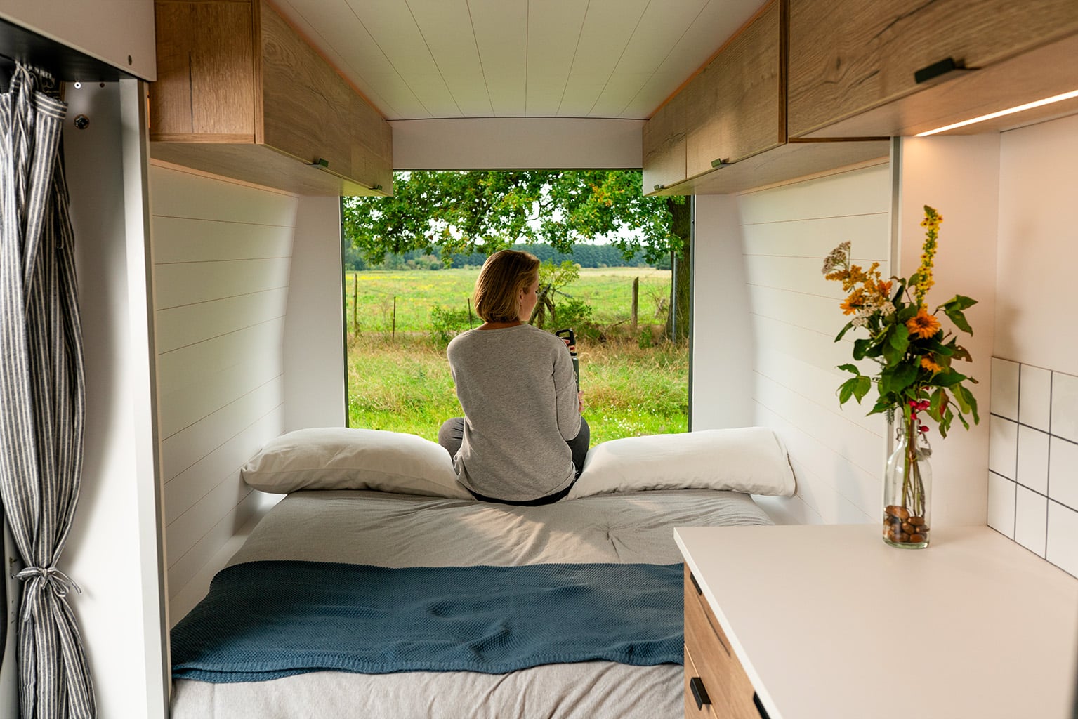 Traveler sitting on the convertible bed of the Biggie Campervan looking out over a field.